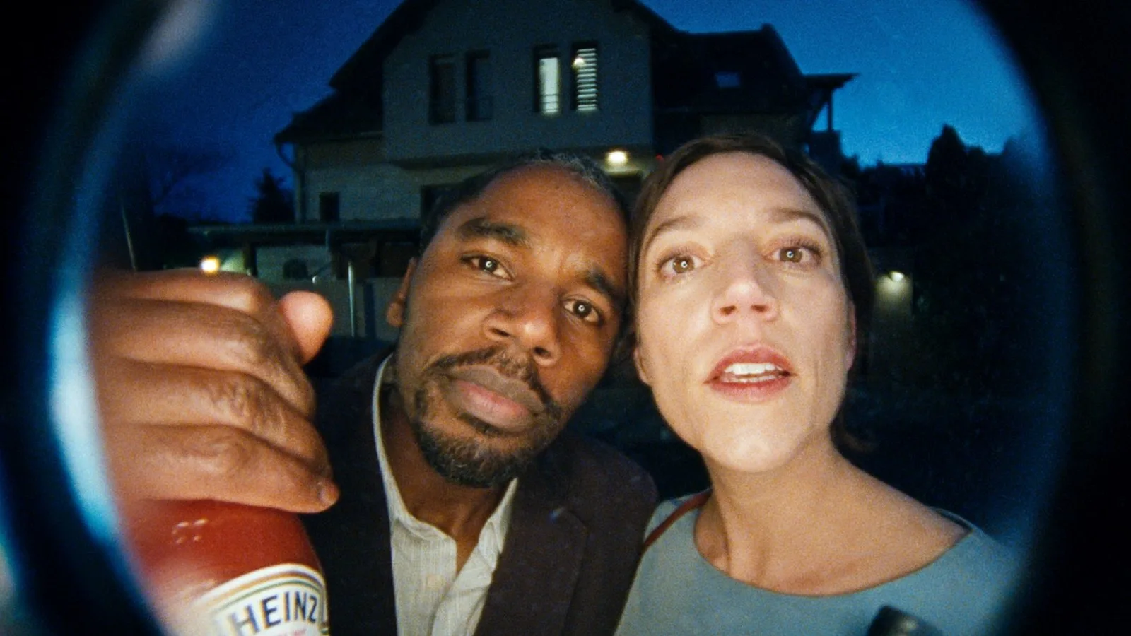 A still from the Heinz campaign — two people seen through a door's peephole at dusk, one holding up a bottle of Heinz.